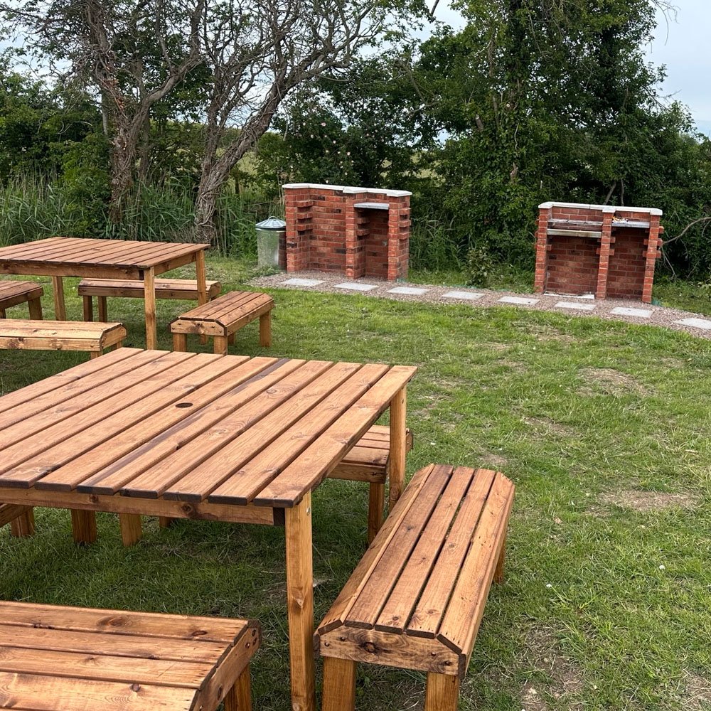 Timber picnic tables and brick-built barbecues in the grass beside the trees
