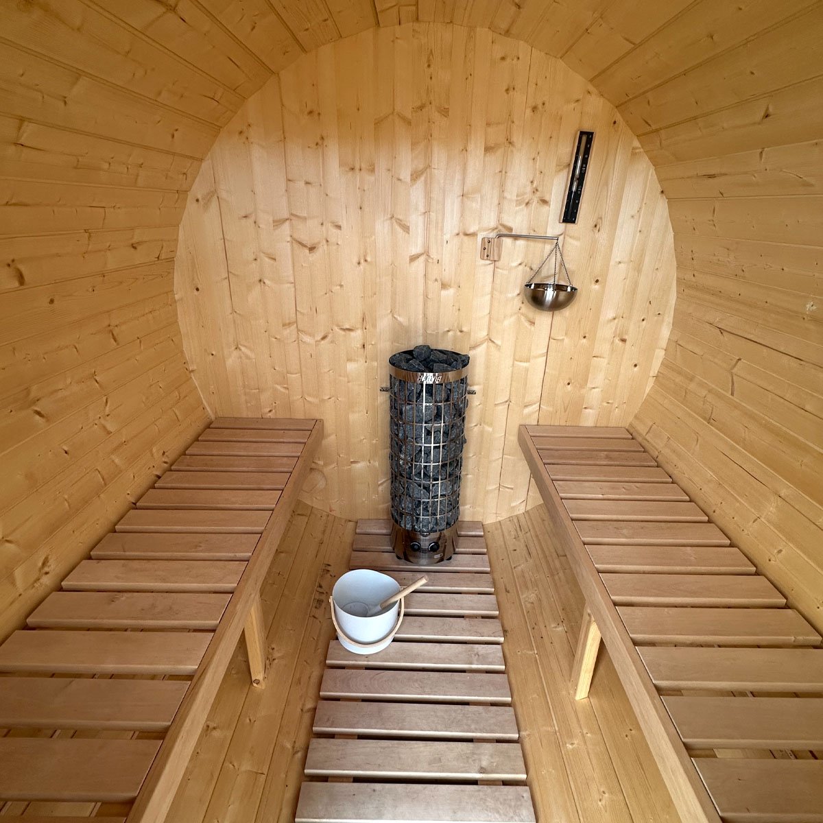 Inside the sauna, with facing timber benches, a stone-filled stove, and a bucket and ladle